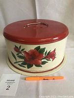 Top and side view of red lid metal cake holder with floral design and metal handle on lid.