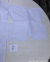 White linen piece with small square cutouts and floral embroidery, scalloped edges with lace trim.
