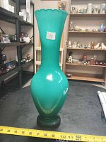 Photo of a tall turquoise green glass vase with scalloped rim and dark base, placed on a black surface. A yellow measuring tape in front shows it is about 15 inches tall. Shelf and other items in background.