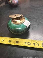 Photo of a green round BMP table lighter with gold-toned metallic top, placed on a black surface with a yellow measuring tape showing about 3.5 inches diameter.