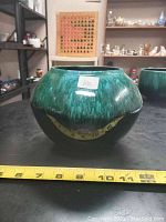 Side view of vintage ceramic bowl/vase showing dark green textured glaze with yellow design and a flat bottom. Tape measure along the base shows approximate size.