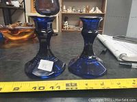Side view of two blue glass candle holders showing flared bases measuring about 4 to 4.5 inches in diameter, placed next to a tape measure for scale.