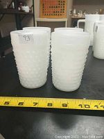 Four vintage white milk glass cups with a hobnail pattern arranged in two pairs, shown with a measuring tape indicating size about 4 inches tall. Shot from the front left angle.