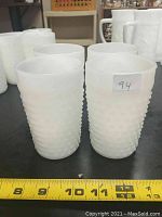 Four vintage milk glass cups with hobnail texture arranged in two rows with measuring tape at base, approx. 4.25 inches tall.