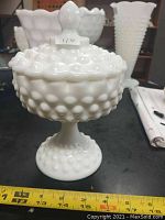 Front angle photo showing white hobnail milk glass covered dish with finial lid and footed base with measuring tape.