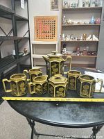 Front view of the entire 7-piece tea set including teapot, sugar bowl, creamer, and mugs arranged on a black table with a tape measure for scale showing about 20 inches width.