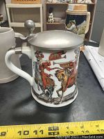 Front and side view of vintage porcelain beer stein decorated with colored mounted musicians, on table with ruler for size reference.