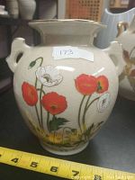 Side view of the ceramic vase showing red and white poppy flowers decoration with green stems, two small handles, and measuring tape indicating size.