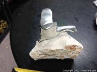 Side view of polished aluminum goose flying sculpture mounted on white stone base, placed on a black surface with a ruler partially visible for scale.