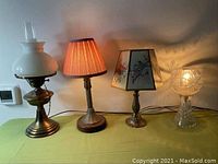 Four small lamps on a yellow-green tablecloth featuring different styles and materials including brass, glass, fabric shades.