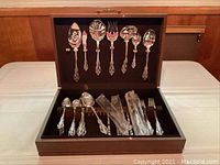 Photo of full silverware set inside open wooden storage box showing hanging serving utensils on the lid and cutlery neatly arranged, some still in original packaging.