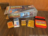 Photo of assorted vintage handheld electronic games and boxed Electronic Battleship game on a wooden surface.