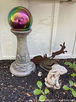 Wide shot showing entire gazing ball on pedestal, rabbit sculpture, and cut iron garden art on outdoor floor against wall.