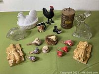 Full view of all items showing piggy banks, rooster figurine, chalkware plaques, miniature Toby mug, milk glass hen, and bird figurines on green tablecloth.