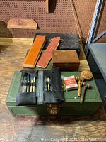Vintage metal cash boxes with two slide rules in black leather cases, two mini wooden gavels, some metal balance weights, a small wooden box, all sitting on a green metal box and a wooden floor.