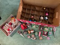 Top view of large box with grid compartments holding various glass ornaments. Additional ornaments and boxed vintage ornament sets arranged on carpet in front of the box.