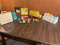 Wide shot showing the full lot of vintage Fisher Price toys on a dark wooden table, including Medical Kit, Cash Register, School Bus, Shape Sorter, and multiple small toy figures.