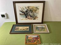 Four framed artworks on green tablecloth - large Old Tree watercolor, horse pasture long watercolor, small English cottage watercolor, Mexican Hacienda chalkware plaque