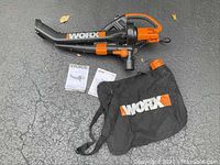 Worx electric blower, collection bag, and two instruction manuals laid out on paved surface showing overall contents of the lot.