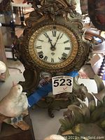 Front view of ornate antique Ansonia clock with Roman numeral face and intricate metal design, alongside bird figurines.