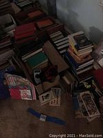 Photo of assorted hardcover and paperback books stacked on the floor with some titles visible.