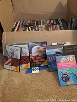 Wide view of multiple DVDs and CDs in a box and some displayed in front, showcasing collection variety.