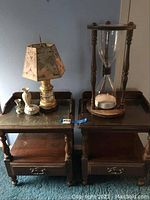 Two wooden end tables with drawers and lower shelves, showing wear. On the left table is a lamp with map-themed shade and small decorative items; on the right table is a large hourglass.