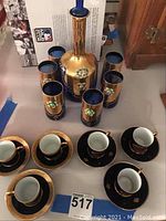 Decorative wine decanter surrounded by six matching tall glasses with floral designs and four espresso cups with saucers arranged at front, all displayed on tabletop.