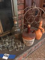Wide view of lot on hearth: two ceramic whiskey jugs - one large Jack Daniels jug with stopper, one smaller jug with figurine on top, metal wine bottle holder, and seven glasses (four square Jack Daniels glasses, three shot glasses).