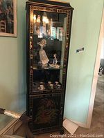 Full view of tall black lacquered wooden cabinet with glass door showing ceramic figurines and tea set inside, surrounded by a frame with gold-painted accents and detailed decoration on the lower door depicting a Japanese outdoor scene with figures and nature.