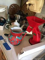 Photo of kitchen counter showing red Moplen grinder, ceramic teapot and pitcher, coffee maker, glass bottle, and woven baskets.