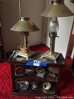 Pair of brass electric lamps and seven compasses displayed on a wooden shelf against a background with red carpet.