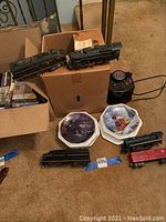 Two Lionel locomotives resting on a cardboard box beside the transformer and decorative plates on floor.