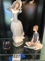 Two Lladro Daisa figurines and a small blue decorative egg on a shelf with a black glossy background. Standing woman in period dress with wide-brimmed hat and corset, and seated ballerina in lavender top are visible.