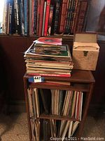 Image showing wooden record case filled with vinyl records and a stack of records on top, with books on shelf behind.