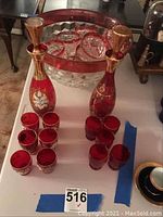 Photo showing entire lot on a table. Includes two red and gold floral decorated decanters, a large clear faceted punch bowl with red rim, and multiple red glasses with gold decoration.