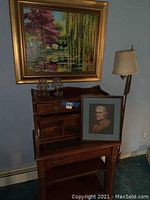 Roll top desk with framed portrait painting resting on it, floor lamp to the right, framed landscape painting above and two metal candle holders on the desk.