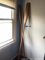Pair of wooden oars leaning against a wall near a window and door, showing full length and shape.