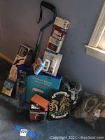 Photo showing white mini blinds in package, aqua-colored cane with black handle, thermometer in sealed packaging, compact round shower stool box, shoes, and other items stacked on the floor near a wall and window.