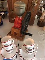 Overall shot showing vintage ELMA red cast-iron coffee grinder and stacked white espresso cups with saucers