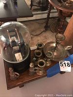 Ticket tape machine under glass dome with visible paper tape reel, accompanied by multiple brass candle holders arranged on a wooden surface.