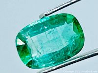 Close-up photo of the natural emerald gemstone held by tweezers, showing the green color, cushion cut and surface reflections.