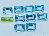 Top-down photo showing 10 emerald cut sky blue topaz stones and 1 rectangular green tourmaline stone arranged on a white background.