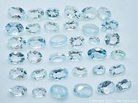 Overview photo showing 36 loose faceted soft blue Aquamarine gemstones, various oval shapes and sizes, laid on white background.