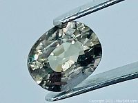 Close-up photo of the oval cut greenish purple natural Tanga sapphire held by tweezers, showing facets and internal inclusions.