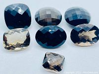 Seven mixed-sized fancy cut Smokey Quartz gemstones arranged in two rows on a white background. The stones have cushion and oval shapes with checkerboard faceting and colors ranging from deep to light smoky brown.