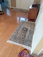 Photo showing two rectangular area rugs laid out on hardwood floor near front door and hallway. The rugs show traditional Persian-style patterns with a central medallion and floral motifs in cream and navy blue tones, fringed ends visible.