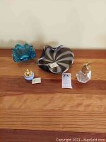 A wooden shelf displaying four items. From left to right: a small blue wavy-edged glass candy dish, a black and white swirled glass candy dish, a small blue cobalt Wedgwood lighter, and a vintage lighter with gold-tone top on a clear glass base.