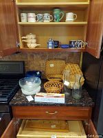 Photo showing mugs on the upper shelves, ceramic soup crocks, blue cracker dish, wicker baskets, glass bowls and glass pitchers on the countertop.