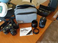 Image showing Olympus OM77AF camera, zoom lens, additional lens tube, black attachment ring, instruction manual, and gray camera bag on a table.